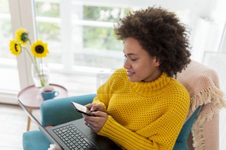 Foto de Hermosa mujer de raza mixta sentada en un sillón, relajándose en casa, utilizando una computadora portátil y escribiendo un mensaje de texto en un teléfono inteligente - Imagen libre de derechos