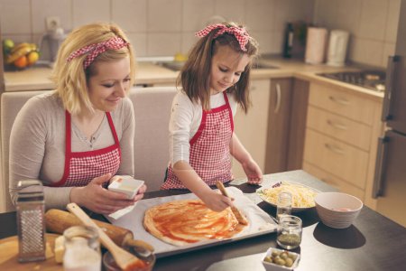 Photo for Mother and daughter in the kitchen making pizza, daughter spreading ketchup on pizza dough with kitchen brush. Focus on the daughter - Royalty Free Image