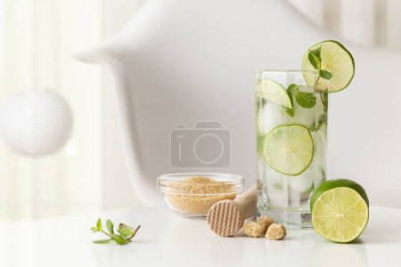 Téléchargez les photos : Cocktail mojito avec beaucoup de glace, rhum blanc, jus de citron et tonique, décoré de tranches de citron vert et de feuilles de menthe sur une table blanche moderne - en image libre de droit