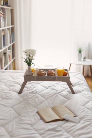 Téléchargez les photos : Plateau de petit déjeuner avec vase de fleurs, jus d'orange, croissants et café placés sur le lit. Focus sur les croissants et le verre - en image libre de droit