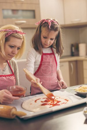 Photo for Mother and daughter in the kitchen making pizza, daughter spreading ketchup on pizza dough with kitchen brush. Focus on the daughter - Royalty Free Image