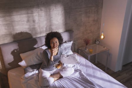 Téléchargez les photos : Vue en angle élevé de belle jeune femme mixte portant un pyjama assis sur le lit, prenant le petit déjeuner et se relaxant à la maison le matin - en image libre de droit