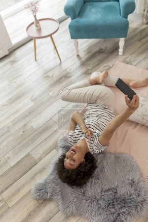 Téléchargez les photos : Vue grand angle d'une belle jeune femme métissée couchée sur le sol du salon, prenant un slefie avec son téléphone intelligent et s'amusant tout en se relaxant à la maison - en image libre de droit
