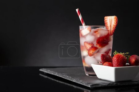 Photo for Glass of cold infused water with fresh strawberry slices and a bowl of fresh strawberries on a black stone tray - Royalty Free Image