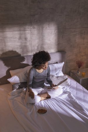 Foto de Vista de ángulo alto de una hermosa joven mestiza con pijamas sentada en la cama, leyendo periódicos, desayunando y disfrutando del tiempo libre en casa - Imagen libre de derechos