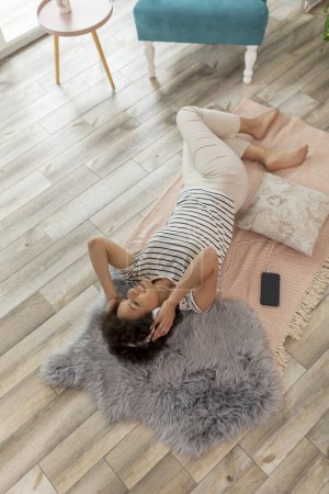 Téléchargez les photos : Vue du dessus de la belle jeune femme mixte couchée sur le sol du salon, portant un casque, écoutant la musique et se relaxant à la maison - en image libre de droit