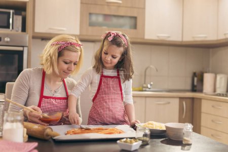 Photo for Mother and daughter in the kitchen making pizza, daughter spreading ketchup on pizza dough with a spoon - Royalty Free Image