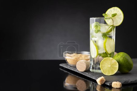 Téléchargez les photos : Cocktail mojito avec beaucoup de glace, rhum blanc, jus de citron et tonique, décoré de tranches de citron vert et de feuilles de menthe sur un plateau en pierre noire - en image libre de droit