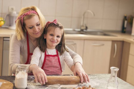 Photo for Mother and daughter baking cookies in the kitchen; little girl using a rolling pin with the help of her mother, kneading cookie dough. Focus on the daughter - Royalty Free Image