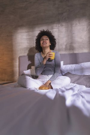 Téléchargez les photos : Belle jeune femme de race mixte portant un pyjama assis sur le lit, prenant un verre de jus d'orange fraîchement pressé pour le petit déjeuner et se détendre à la maison le matin - en image libre de droit