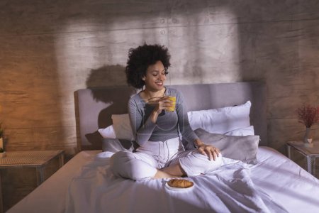 Foto de Hermosa joven mujer de raza mixta con pijama sentado en la cama, desayunando y relajándose en casa por la mañana - Imagen libre de derechos