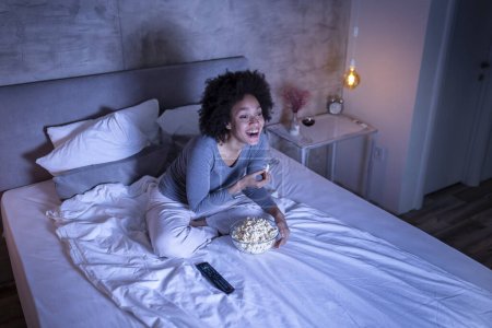 Téléchargez les photos : Belle femme de race mixte portant un pyjama assis sur le lit, mangeant du pop-corn et regardant un film comique à la télévision, se détendre à la maison tard dans la nuit - en image libre de droit