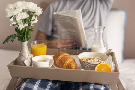 Téléchargez les photos : Homme en pyjama, lisant les journaux et prenant le petit déjeuner au lit. Focus sélectif sur le croissant et le vase - en image libre de droit