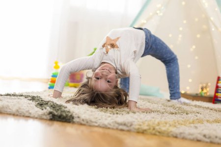 Photo for Cheerful little girl doing backbend, playing in her playroom and having fun - Royalty Free Image