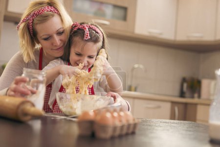 Foto de Hermosa niña amasando masa con la ayuda de sus madres, divirtiéndose en la cocina - Imagen libre de derechos