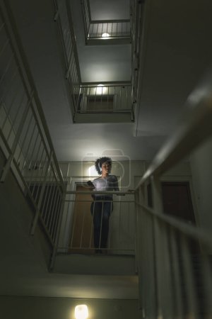 Photo for Young woman standing on a low light staircase in a building hallway, listening to the music; woman wearing a headset and choosing a song from a smart phone playlist - Royalty Free Image