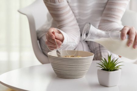 Foto de Detalle de las manos femeninas vertiendo leche en un tazón de cereales; mujer desayunando. Concéntrate en el cereal y la punta de la botella - Imagen libre de derechos