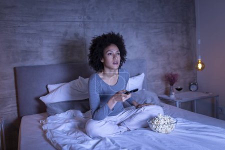 Téléchargez les photos : Belle jeune femme de race mixte portant un pyjama assis sur le lit, mangeant du pop-corn et changeant de chaînes à la télévision avec une télécommande - en image libre de droit