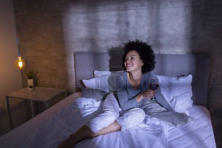 Foto de Mujer joven de raza mixta con pijamas sentada en la cama, tomando una copa de vino y relajándose en casa después de un día ajetreado. - Imagen libre de derechos