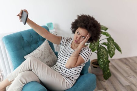Foto de Hermosa joven mujer de raza mixta sentada en un sillón, tomando una selfie con su teléfono inteligente y divirtiéndose mientras se relaja en casa - Imagen libre de derechos