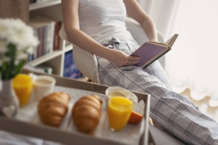 Téléchargez les photos : Femme en pyjama, assise sur une chaise à côté du lit, lisant un livre. Plateau de petit déjeuner au premier plan. Focus sélectif sur le haut du livre - en image libre de droit