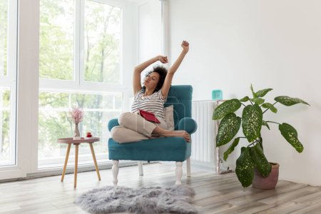 Photo for Beautiful young mixed race woman stretching while sitting in an armchair by the window, reading a book and enjoying leisure time at home - Royalty Free Image