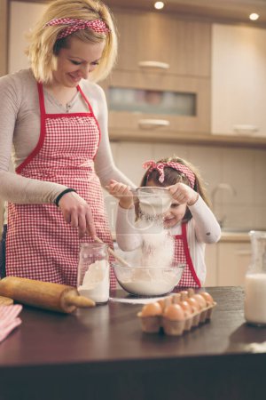 Foto de Madre e hija horneando masa; hija sembrando harina a través del tamiz en un tazón de amasar. Enfoque selectivo en el tamiz y la harina - Imagen libre de derechos