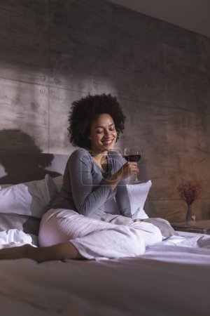 Photo for Woman wearing pajamas sitting on the bed with eyes closed and smiling, holding a wineglass and enjoying leisure time at home - Royalty Free Image