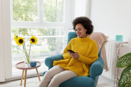 Téléchargez les photos : Belle jeune femme métisse assise dans un fauteuil, relaxante à la maison, tapant un message texte à l'aide d'un téléphone intelligent - en image libre de droit