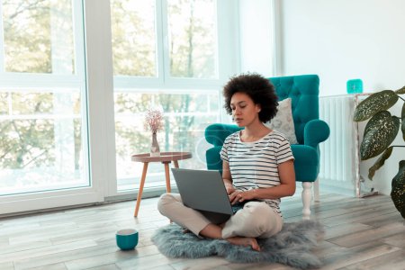 Foto de Freelancer sentada en el piso de la sala de estar, trabajando en una computadora portátil y bebiendo café; mujer trabajando remotamente desde casa - Imagen libre de derechos