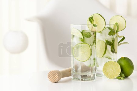 Two mojito cocktails with lots of ice, white rum, lemon juice and tonic, decorated with lime slices and mint leaves on a modern white table.