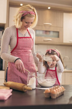 Téléchargez les photos : Mère et fille semant de la farine à travers le tamis dans un bol. Mère apprend à sa fille à pétrir une pâte. Focus sur la fille - en image libre de droit