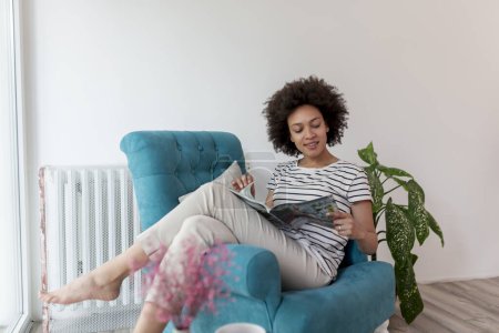 Téléchargez les photos : Belle jeune femme métissée assise dans un fauteuil près de la fenêtre, lisant un magazine et profitant de loisirs à la maison - en image libre de droit