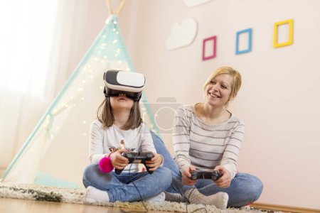 Foto de Madre e hija sentadas en el suelo en una sala de juegos, jugando al juego 3D VR y divirtiéndose. Concéntrate en la hija - Imagen libre de derechos