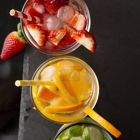 Foto de Vista superior de vasos de agua infundida con lima fresca, pepino y hojas de menta, naranja y fresas. Concéntrate en el vaso de naranja - Imagen libre de derechos