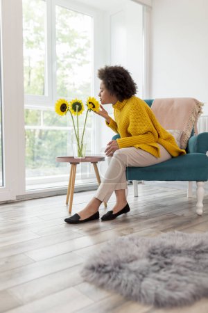 Téléchargez les photos : Belle jeune femme métissée assise dans un fauteuil près de la fenêtre, relaxante à la maison, sentant le tournesol posé dans un vase à côté d'elle - en image libre de droit