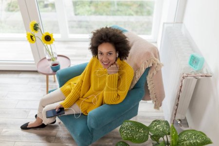 Foto de Vista de ángulo alto de la hermosa joven mestiza sentada en un sillón, sosteniendo un teléfono inteligente, escuchando la música y relajándose en casa - Imagen libre de derechos
