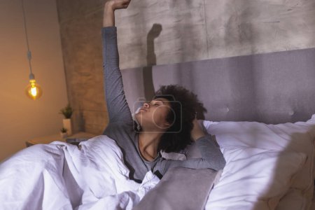 Téléchargez les photos : Belle jeune femme de race mixte portant un pyjama couché dans le lit le matin, étirant et se relaxant à la maison après le réveil - en image libre de droit