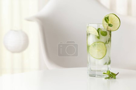 Foto de Cóctel Mojito con mucho hielo, ron blanco, zumo de limón y tónico, decorado con rodajas de lima y hojas de menta en una moderna mesa blanca - Imagen libre de derechos