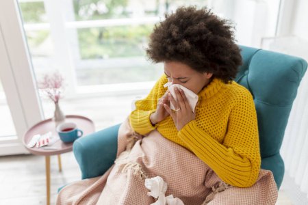 Foto de Retrato de una mujer enferma sentada en un sillón cubierto con manta, con gripe y fiebre, sonando la nariz en un papel - Imagen libre de derechos