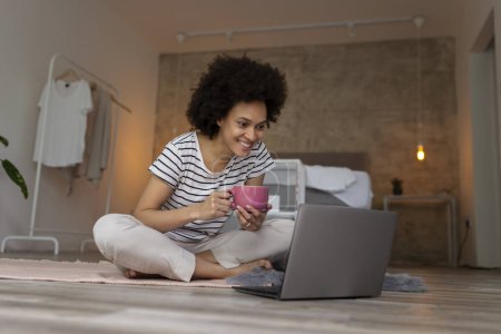 Téléchargez les photos : Belle jeune femme métisse assise sur le sol de la chambre à coucher, ayant un appel vidéo sur un ordinateur portable et buvant du café - en image libre de droit