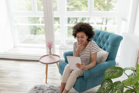 Photo for Beautiful young mixed race woman sitting in an armchair, having a video call using tablet computer; woman enjoying her leisure time activities at home - Royalty Free Image