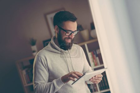 Téléchargez les photos : Beau jeune homme travaillant sur un ordinateur tablette tout en se tenant à côté d'une fenêtre de bureau à la maison - en image libre de droit