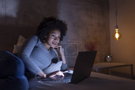 Foto de Hermosa joven mujer de raza mixta acostada en la cama por la noche, teniendo una videollamada en una computadora portátil - Imagen libre de derechos