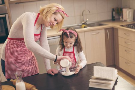 Foto de Madre e hija horneando masa en la cocina; madre agregando harina de un frasco mientras la hija se agita con una cuchara de cocina. Concéntrate en la hija - Imagen libre de derechos