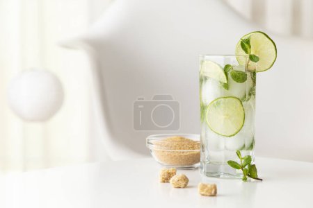 Photo for Mojito cocktail with lots of ice, white rum, lemon juice and tonic, decorated with lime slices and mint leaves on a modern white table - Royalty Free Image