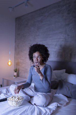 Foto de Hermosa mujer de raza mixta sentada en la cama, bebiendo vino, comiendo palomitas de maíz y viendo televisión - Imagen libre de derechos