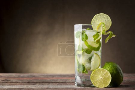 Photo for Mojito cocktail with lots of ice, white rum, lemon juice and tonic, decorated with lime slices and mint leaves on a rustic wooden table - Royalty Free Image