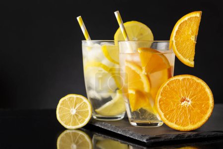 Photo for Glasses of cold infused water with fresh lemon and orange slices on a black stone tray. Focus on the orange infused water - Royalty Free Image