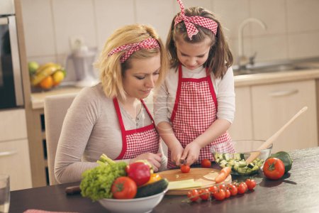 Téléchargez les photos : Mère et fille coupant des légumes et faisant de la salade ; petite fille coupant des tomates cerises, aidant sa mère dans la cuisine - en image libre de droit